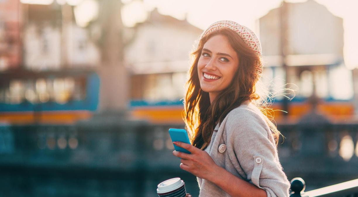 Mujer sonriendo con un móvil en una mano y café en la otra