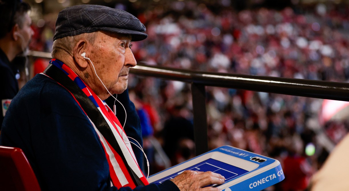 Un aficionado con bufanda del Atlético de Madrid y una tablet y cascos para seguir el partido con el audio y el tacto